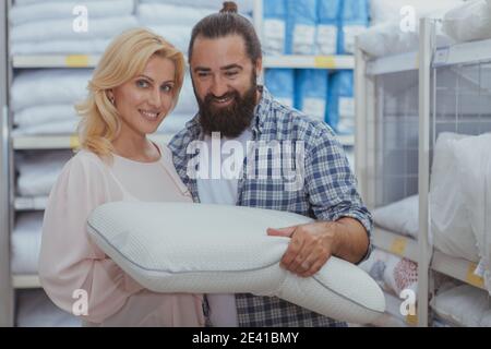 Concetto di prodotti ortopedici. Bella donna matura sorridente alla macchina fotografica, mentre lo shopping per cuscini ortopedici con suo marito. Coppia che compra la biancheria da letto Foto Stock