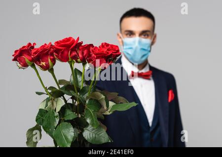 elegante uomo ispanico in maschera medica con rose rosse isolate su sfondo grigio, sfocato Foto Stock