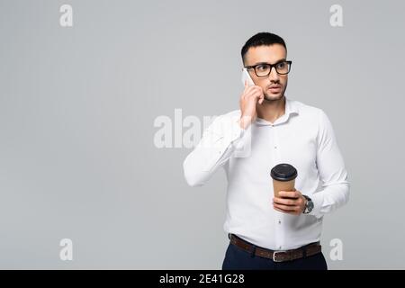 uomo d'affari ispanico in occhiali che parla su smartphone mentre si tiene il caffè per essere isolato su grigio Foto Stock