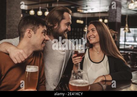 Felice uomo bearded abbracciando i suoi amici, mentre bevendo birra al pub locale. Gruppo di amici allegri che abbracciano, parlano e celebrano il Tog Foto Stock