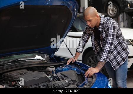 Giovane uomo africano che esamina il motore della sua auto sportiva al garage, guardando sotto il cofano Foto Stock