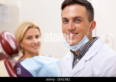 Bel giovane dentista maschio che sorride alla telecamera, la sua paziente femmina felice sullo sfondo. Dentista professionista che si diverse a lavorare presso la sua clinica. De Foto Stock