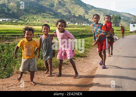 Manandoana, Madagascar - 26 aprile 2019: Gruppo di bambini malgascio sconosciuti che corrono su strada accanto al campo di riso, piccole colline sullo sfondo, persone a Madag Foto Stock