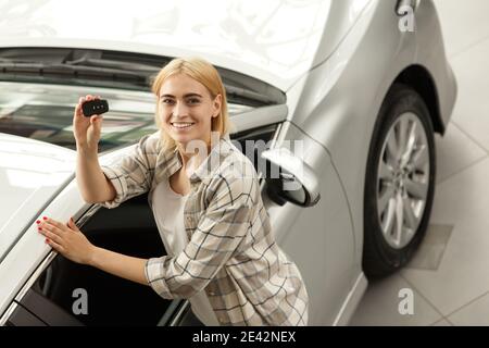 Scatto orizzontale di una bella giovane e felice driver femmina sorridente tenendo le chiavi dell'automobile alla sua nuova automobile. Donna allegra che posa vicino a lei ha appena comprato un Foto Stock