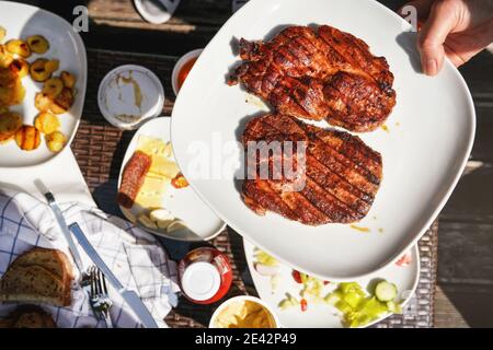Due bistecche di lombo di maiale appena grigliate su piatto bianco tenuto a mano sopra il tavolo da giardino, con più cibo dalla griglia. Foto Stock