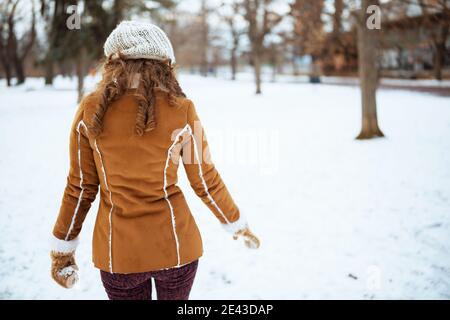 Visto da dietro donna di mezza età con i mitens in un cappello lavorato a maglia e cappotto di pelle di pecora che cammina fuori nel parco della città in inverno. Foto Stock