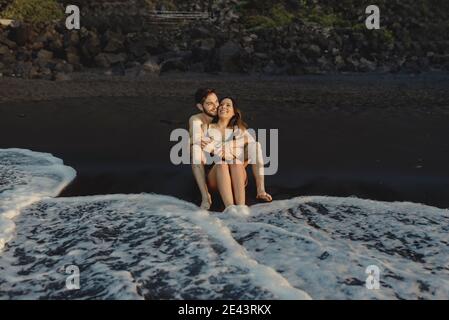 Corpo pieno di felice giovane e donna che abbraccia e ride felicemente mentre si siede sulla spiaggia di sabbia vicino alle onde del mare durante il viaggio di nozze a Tenerife isla Foto Stock