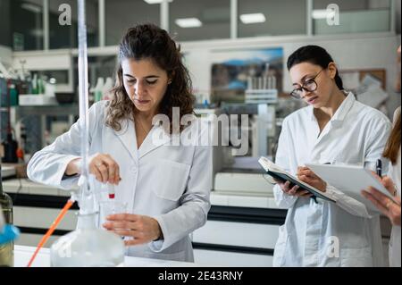 Scienziato femminile concentrato che conduce esperimenti chimici con liquido in vetro matracci mentre colleghi scrivono i risultati durante la ricerca in labo Foto Stock