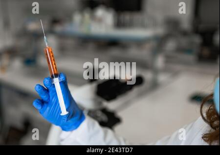 Crop scienziato femminile in maschera medica e guanti di protezione tenendo siringa con campione di vaccino mentre si lavora in un laboratorio moderno Foto Stock