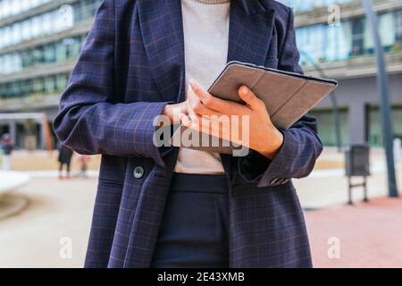 Femminile non riconoscibile dal taglio ad angolo basso con elegante tuta da lettura formale tablet digitale in posizione di fronte a un moderno edificio di uffici in centro Foto Stock