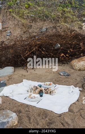 Coperta bianca con cibo e decorazioni allestite per un picnic romantico sulla spiaggia di sabbia con pietre in riva al mare Foto Stock