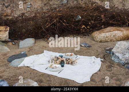 Coperta bianca con cibo e decorazioni allestite per un picnic romantico sulla spiaggia di sabbia con pietre in riva al mare Foto Stock