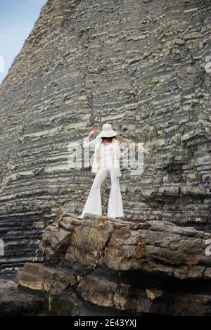 Intera lunghezza di donna asiatica irriconoscibile in elegante vestito bianco e cappello che tiene bouquet di pampas secco pianta mentre in piedi contro montagna rocciosa Foto Stock