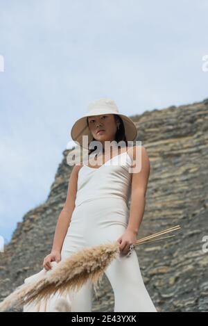 Dal basso di giovane donna asiatica in elegante vestito bianco e cappello che tiene bouquet di piante essiccate mentre in piedi contro montagna rocciosa Foto Stock