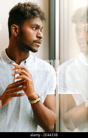 Serio giovane nero sopportato maschio con capelli ricci in casual camicia che guarda lontano pensieroso mentre si è in piedi vicino alla finestra Foto Stock