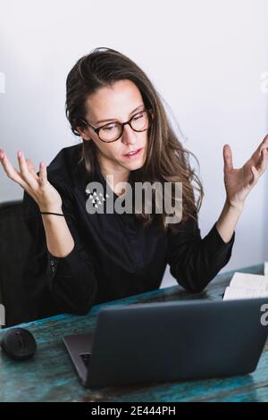 Turbato giovane femmina in abito formale facendo videochiamata su computer portatile mentre si lavora in remoto a casa su un tavolo di legno shabby Foto Stock