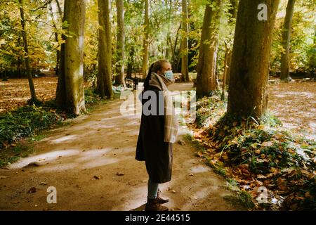 Vista laterale della maschera facciale femmina in piedi lungo il percorso in foresta durante il fine settimana passeggiata in autunno Foto Stock