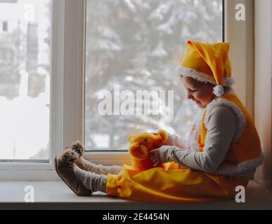 Piccolo bambino ragazzo carino sedersi alla finestra e giocare con il giocattolo elefante, indossando elfo gnome giallo costime con divertente cappello, gilet, pantaloni e pantofole, famiglia invernale Foto Stock