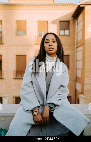 Giovane donna sicura che indossa un cappotto blu alla moda e che guarda macchina fotografica seduta su recinzione in pietra in città Foto Stock