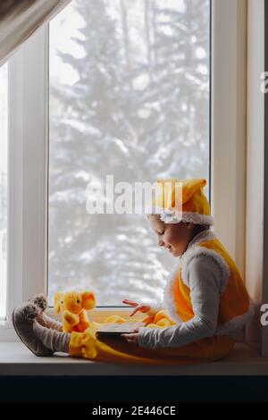 Piccolo ragazzo sorridente in abito giallo gnome elfo sedersi alla finestra con vista invernale e libro di lettura per il giocattolo elefante, famiglia magica tempo di casa invernale Foto Stock