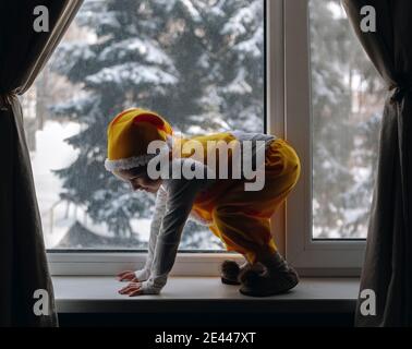 Bambino 4 anni giocare alla finestra con vista invernale indossare costume giallo di ghomo, elfo con gilet, pantaloni e cappello divertente Foto Stock