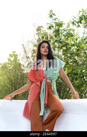 Modello giovane donna etnico sicuro e attraente che indossa un abito colorato ed elegante guardando la macchina fotografica mentre si sta in piedi sulla terrazza contro le piante verdi Foto Stock