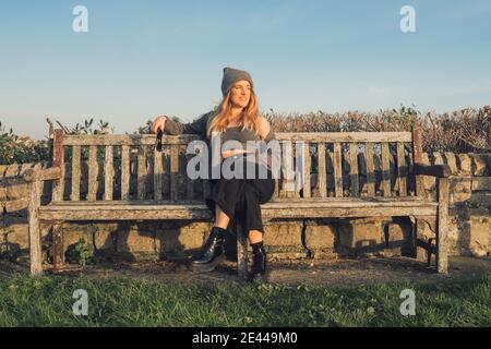 Giovane donna allegra in abiti eleganti e cappelli seduti su una panchina di legno intemperie e godendo di una soleggiata giornata autunnale durante viaggiare nella natura Foto Stock