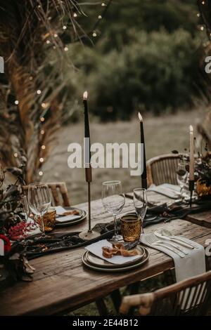 Tavolo di legno servito con bicchieri e piatti e decorato con candele bruciate in giardino per la celebrazione del matrimonio Foto Stock
