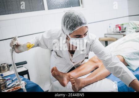 Medico che indossa una maschera, un cappuccio, guanti e una uniforme che tiene uno strumento a ultrasuoni per un paziente che si trova su una barella in un ospedale Foto Stock