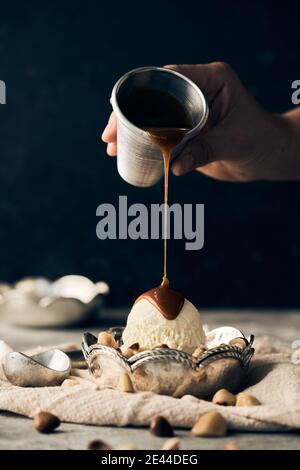 Persona di raccolto irriconoscibile che versa il caramello dolce che topping su ghiaccio di vaniglia paletta per crema in recipiente posto sul tavolo con noci sparse Foto Stock
