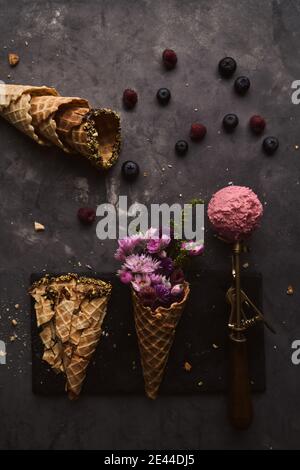 Da sopra di scoop di gelato rosa messo sopra tavolo con cono di wafer con fiori e bacche sparse su sfondo scuro Foto Stock