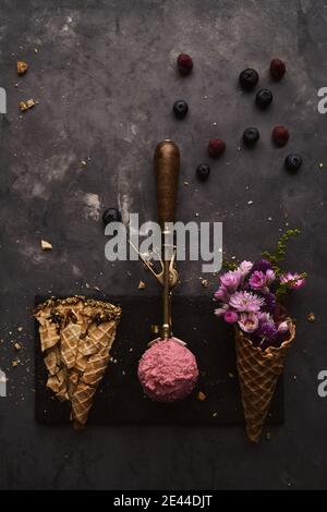 Da sopra di scoop di gelato rosa messo sopra tavolo con cono di wafer con fiori e bacche sparse su sfondo scuro Foto Stock