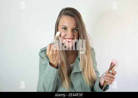 Felice giovane donna sicura con lunghi capelli biondi e rosso labbra in elegante giacca di pelle sorridente e guardando la fotocamera durante l'applicazione della fondazione con b Foto Stock