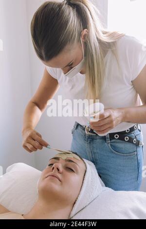 Cosmetician applicando maschera di alginato sul viso di anonima giovane femmina cliente sdraiato sul divano con gli occhi chiusi Foto Stock