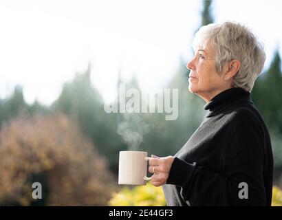 Donna anziana vestita di capelli bianchi e neri bevendo una tazza di caffè dalla finestra a casa in inverno Foto Stock