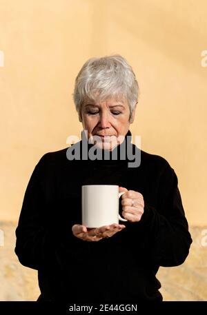 Donna anziana in vestito nero con capelli bianchi bere una tazza di caffè in inverno giorno di sole Foto Stock
