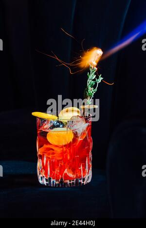 Un bicchiere di fresco cocktail Negroni con una fetta di arancia guarnita con fetta di arancia e rogo di rosmarino in poltrona in velluto Foto Stock