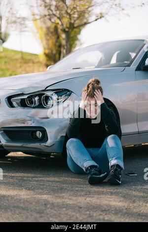 Intera lunghezza di giovane driver triste in abiti casual seduto sulla strada vicino a un'auto costosa danneggiata e piangendo dopo incidente Foto Stock