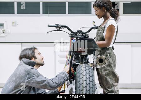 Diversi meccanici maschili e femminili nell'abbigliamento da lavoro che fissano la motocicletta insieme in garage luminoso Foto Stock