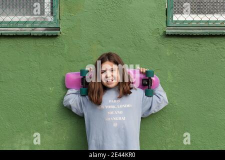 Felice ragazza adolescente in abito casual e sneakers in piedi vicino edificio verde invecchiato con skateboard dietro la testa dopo la guida Foto Stock