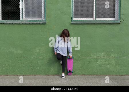 Corpo pieno di ragazza adolescente felice in abito casual e. sneakers in piedi vicino a un edificio verde vecchio con skateboard dietro la testa dopo la guida Foto Stock