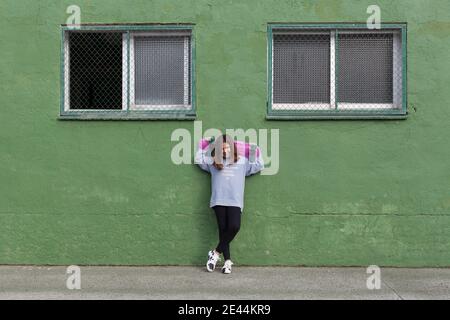 Corpo pieno di ragazza adolescente felice in abito casual e. sneakers in piedi vicino a un edificio verde vecchio con skateboard dietro la testa dopo la guida Foto Stock