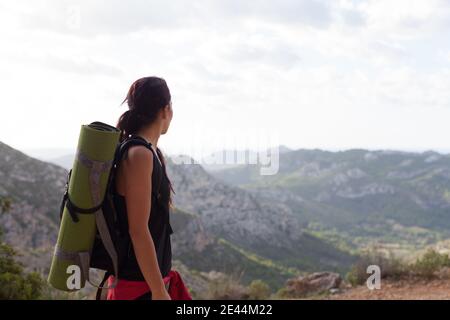 Vista laterale di una giovane escursionista irriconoscibile in activewear e. zaino ammirando il pittoresco paesaggio di mare circondato da montagne verdi in piedi Foto Stock