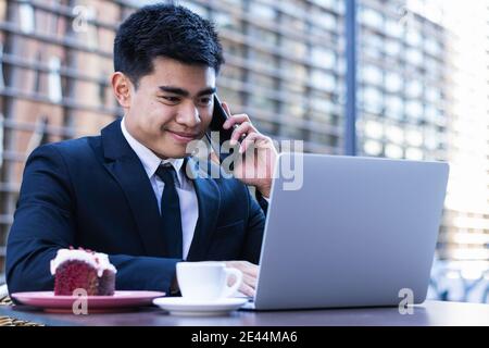 Sorridente imprenditore asiatico seduto al tavolo in un caffè di strada e lavorare in remoto mentre si digita sul computer portatile e si parla smartphone Foto Stock