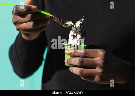 Crop irriconoscibile africano americano maschio in maglione nero mangiare delizioso gelato alla vaniglia da asporto con condimento al cioccolato con cucchiaio contro blu Foto Stock
