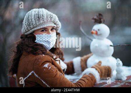 felice elegante donna con mittens e maschera medica in un cappello a maglia e camice di pelle di pecora che fa un pupazzo di neve fuori nel parco della città in inverno. Foto Stock