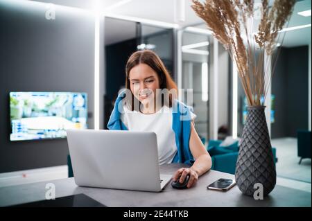 Donna sorridente seduto in cucina e utilizzando il computer portatile. Lavoro da casa in quarantena. Isolamento di sé di allontanamento sociale. Foto Stock