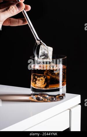 Crop barman anonimo con pinze di metallo che versano cubetti di ghiaccio in Bicchiere mentre si prepara un cocktail vecchio stile con whisky Foto Stock