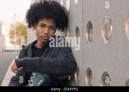 Serio adolescente africano americano hipster maschile con capelli curly cercando alla macchina fotografica mentre si siede vicino al muro di cemento sulla strada urbana Foto Stock