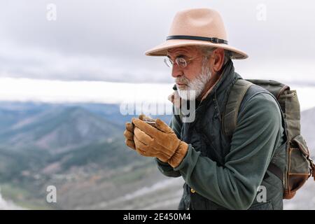 Anziano escursionista maschile in outerwear in piedi in montagna in inverno E bere caffè caldo in tazza durante la vacanza a Caceres Foto Stock